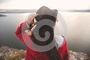 Traveler girl in hat and with windy hair standing in evening sunlight on top of rock mountain, enjoying beautiful sunset view on