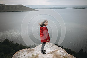 Traveler girl in red raincoat and with windy hair standing on top of rock mountain with beautiful view on river. Young hipster