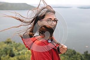 Traveler girl in red raincoat and with windy hair standing on top of rock mountain, enjoying beautiful view on river. Young