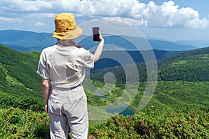 The traveler in a hat and backpack is resting in nature. A tourist girl photographs the panorama of the mountains and the lake.