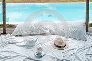 Traveler hat, hot tea cup and teapot on white bed against Beautiful ocean view background, Tourists relaxing in tropical resort