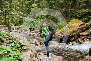 Traveler hiker with backpack enjoys landscape of mountain river in Carpathian forest. Young woman walks on rocks