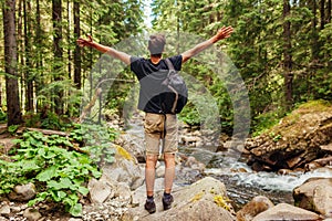 Traveler hiker with backpack raising arms by mountain river in Carpathian forest feeling happy. Trip to summer Ukraine