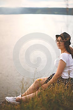 Traveler hipster girl in hat and windy hair sitting on top of rock mountain in grass, enjoying beautiful sunset view on river.
