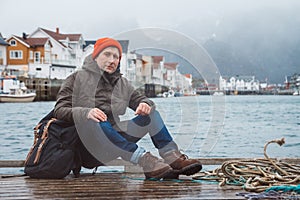 Traveler man with backpack sitting on wooden pier on the background of lake and mountain houses. Space for your text message or