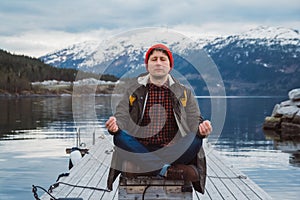 Traveler man in a meditative position sitting on a wooden pier on the background of a mountain and a lake. Space for your text