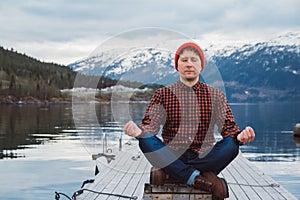 Traveler man in a meditative position sitting on a wooden pier on the background of a mountain and a lake. Space for your text