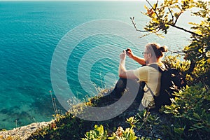 Traveler man sits on shore and takes pictures of sea on cell phone camera during sunset