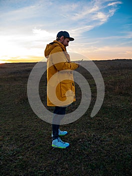 Traveler man in yellow coat with smartphone walking on a hill sunset sky side view. Authentic male tourist with gadget