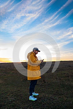 Traveler man in yellow coat with smartphone walking on a hill sunset sky side view. Authentic male tourist with gadget