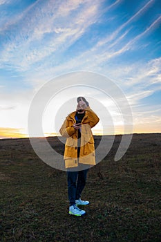 Traveler man in yellow coat with smartphone walking on a hill sunset sky side view. Authentic male tourist with gadget