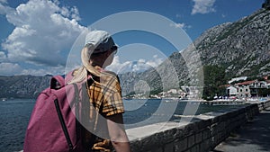 Traveler with a pink backpack walks along the road by the sea, against the background of the old city