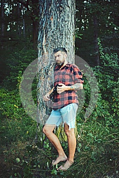 Traveler in plaid shirt and jean shorts travel barefoot. Bearded man with shovel in forest. Hipster with long beard on