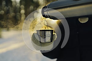 Traveler pours hot tea from a thermos into a mug, walking in snowy frozen winter forest