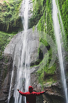 Traveler in red jacket raised hand, at Madakaripura waterfall in Indonesia. Travel lifestyle and success