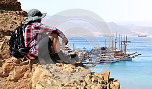 Traveler sits on the rock edge and looks at harbor. View from cliff on Eilat Red sea with the sailing vessels moored in the bay