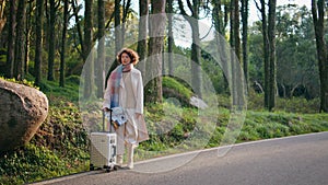 Traveler standing roadside suitcase at autumnal forest. Woman waiting journey