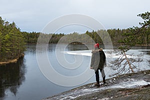 Traveler standing on rock. Cloudy dramatic winter sky. Cold weather, snowy lakesides with green fir forest. Travel concept.