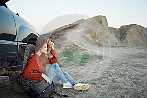 a traveler in a sweater and jeans sits on the ground with a backpack in her hand near a car in the mountains