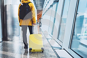 Traveler tourist in bright jacket with yellow suitcase backpack at airport on background large window blue sky, man waiting