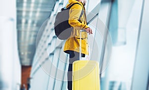 Traveler tourist in bright jacket with yellow suitcase backpack at airport on background large window blue sky, man waiting