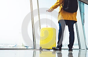 Traveler tourist hold yellow suitcase backpack is standing at airport on background large window, man in bright jacket waiting