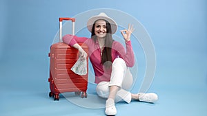 Traveler tourist woman in casual clothes and white hat holding money and sitting at the red suitcase isolated on blue background