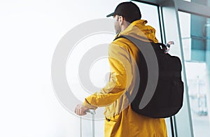 Traveler tourist with yellow backpack is standing at airport on background large window, man in bright jacket waiting in departure