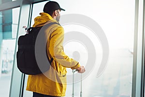 Traveler tourist with yellow backpack is standing at airport on background large window, man in bright jacket waiting in departure