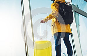 Traveler tourist with yellow suitcase backpack is standing at airport on background large window, man in bright jacket waiting