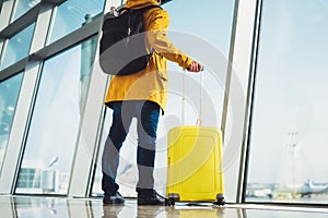 Traveler tourist with yellow suitcase backpack is standing at airport on background large window, man in bright jacket waiting