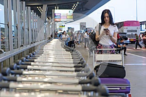 Traveler woman in airport terminal using mobile smartphone with luggage and bag on airport trolley cart with busy airport
