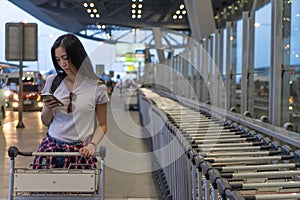 Traveler woman in airport terminal using mobile smartphone with luggage and bag on airport trolley cart..Young asian girl tourist