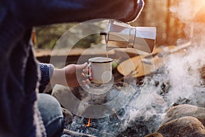 Traveler woman in blue sweater pours itself hot beverage in mountains near to bonfire, relaxing after trekking.