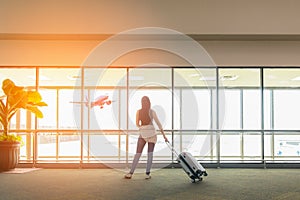 Traveler woman standing and hand holding luggage bag see the airplane at the airport glass window. Tourist waiting in hall airpla