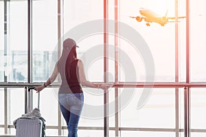 Traveler woman standing and hand holding luggage bag see the airplane at the airport glass window.