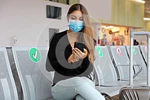 Traveler woman with surgical mask using smart phone and sitting respecting social distancing at the airport lounge