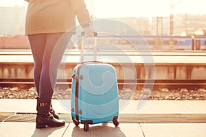 Traveler woman waiting at railway station. Travel by train. Girl standing with blue luggage suitcase in morning. Journey concept.