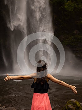 Traveler woman wearing pink dress at waterfall. Excited woman raising arms in front of waterfall. Travel lifestyle. View from back