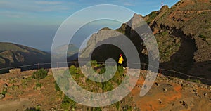 A traveler in a yellow jacket, hat and backpack walks on a paradise island. Active young woman climbs on top of cliff