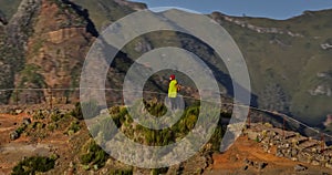 A traveler in a yellow jacket, hat and backpack walks on a paradise island. Active young woman climbs on top of cliff