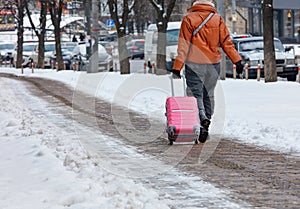 A traveling woman walks along a winter paved pavement cleared of snow, carrying a suitcase on wheels. Copy space