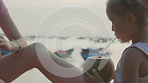 Travelers mother and daughter sits at seafront and drinks coconats in trip