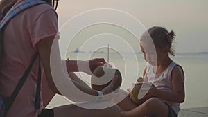 Travelers mother and daughter sits at seafront and drinks coconats in trip
