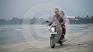 Travelers ride motorbike along sandy sea beach at dusk. Dog running chasing bike. Man, woman drive motorcycle on