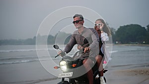 Travelers ride motorbike along sandy sea beach at dusk. Man, woman drive motorcycle on tropical island coastline