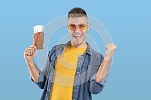 Traveling abroad concept. Excited young guy holding passport with tickets and gesturing YES over blue studio background