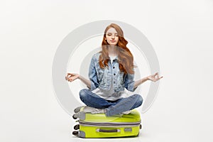 Traveling concept. Young pretty ginger woman meditating in lotus pose on the luggage valise. Isolated on white.