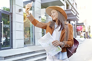 Traveling in Europe. Young happy female traveler or tourist wearing hat and backpack holding map, making selfie while