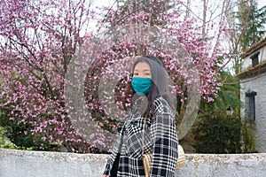 Traveling in virus times - young happy and beautiful Asian Korean woman in face mask against covid19 enjoying sakura blooming tree